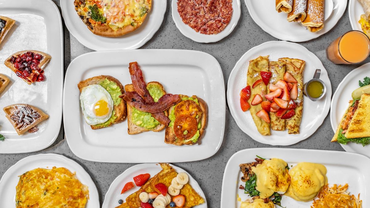 A top-down flat lay view of a diverse breakfast and brunch spread featuring avocado toast, eggs, waffles, crepes, and fresh juice.