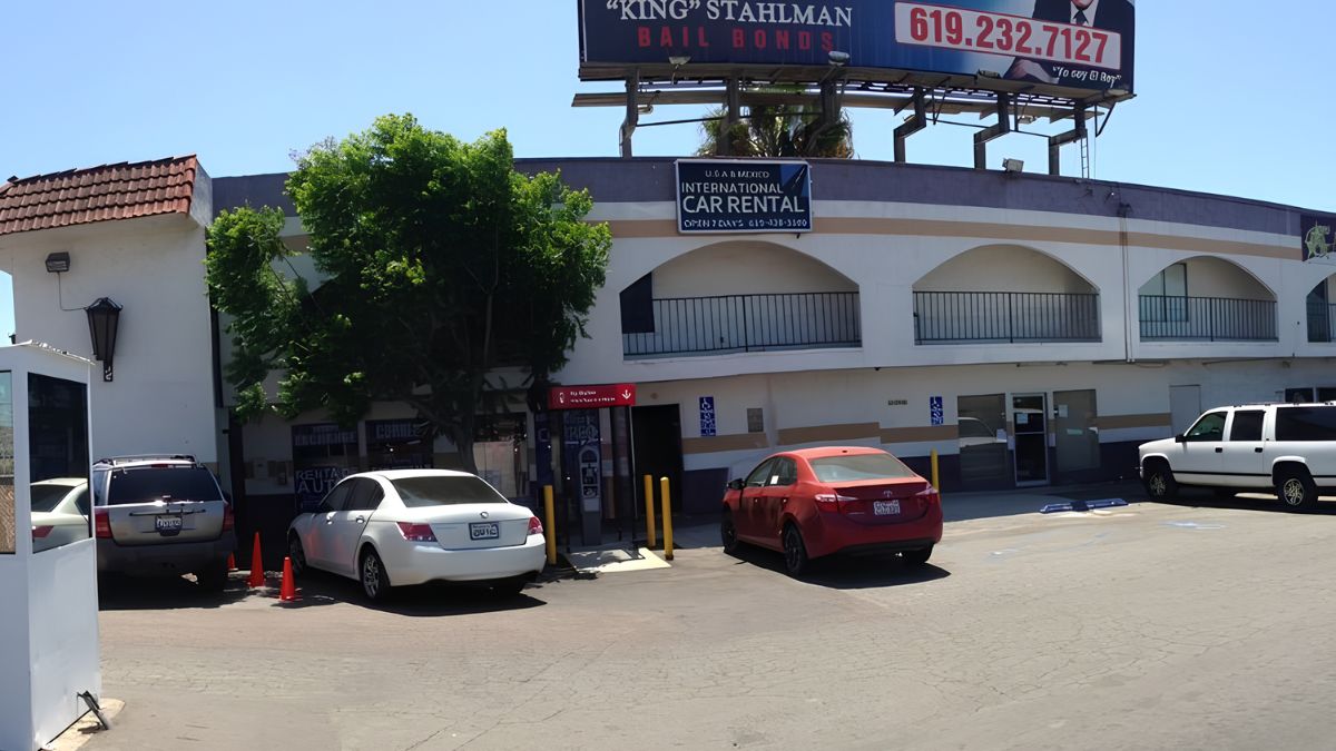 Exterior view of a two-story stucco commercial building on a sunny day, housing "INTERNATIONAL CAR RENTAL" in San Ysidro. Several vehicles, including a white sedan, a red sedan, and an SUV, are parked in the asphalt lot in front. A large billboard for "KING" STUHLMAN BAIL BONDS is visible on the roof.
