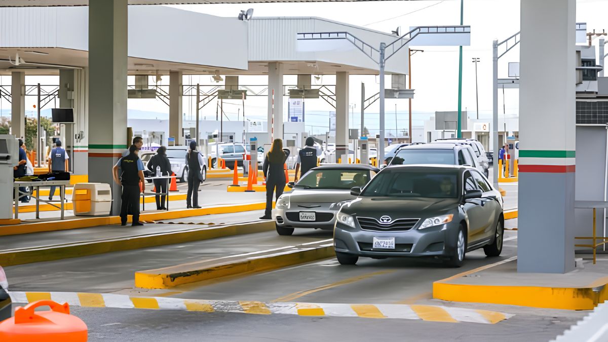 A daytime photograph of a border crossing inspection lane. A gray Toyota sedan approaches the inspection point where two U.S. Customs and Border Protection officers in dark uniforms are stationed. Another agent stands to the left near a group of people.