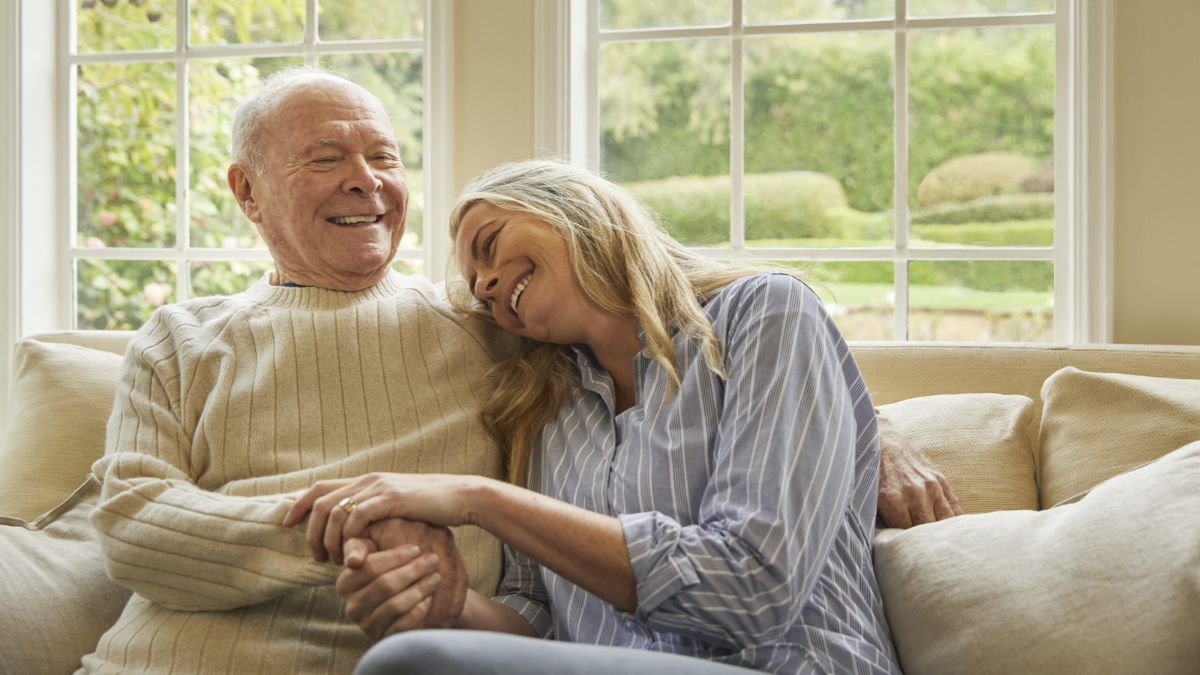 An elderly man smiling happily while sitting on a couch with a woman holding his hand and resting on his shoulder
