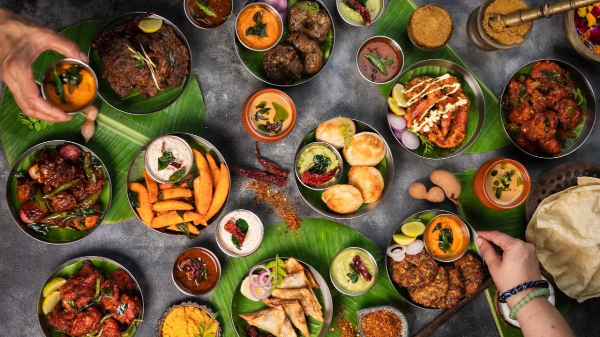 A vibrant, top-down flat lay view of a large spread of traditional South Indian dishes served on banana leaves, featuring curries, fried snacks, chutneys, and breads, with hands reaching in.