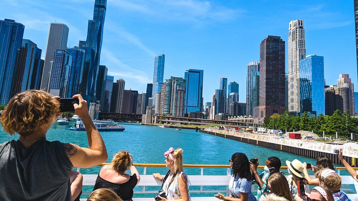 A group of diverse tourists, seen from the back and side, stand on the deck of a boat, facing the Chicago River. In the foreground, a person with long curly hair holds a smartphone up to take a picture of the city. Other passengers are also using their phones or cameras. Beyond the river, a dense cluster of tall skyscrapers under a clear blue sky forms the iconic Chicago skyline.