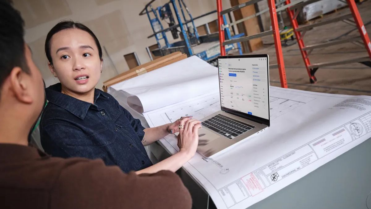A female construction project manager, wearing a denim shirt, is pointing at a laptop screen while explaining details to a male colleague. The laptop displays project management software. They are standing at a table covered with rolled-up architectural blueprints inside a building under construction with scaffolding and ladders in the background.