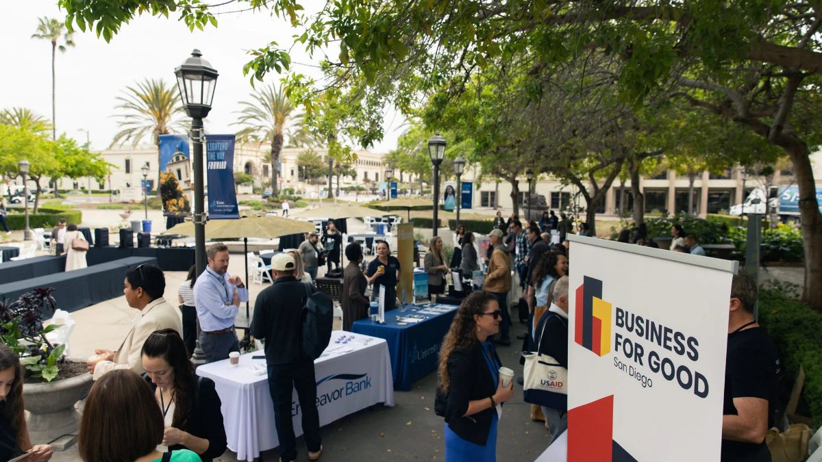 Attendees network among exhibitor booths at the Business for Good San Diego outdoor event, held on a sunny plaza with Mission-style buildings in the background.