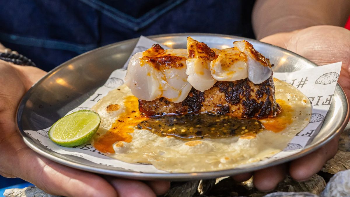 A person holding a metal plate featuring a taco topped with seafood, spicy paste, salsa, and a lime wedge