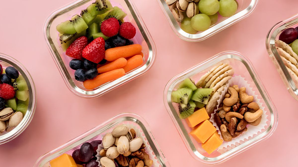 Glass containers filled with healthy snacks like fresh fruits, nuts, and crackers on a pink background.