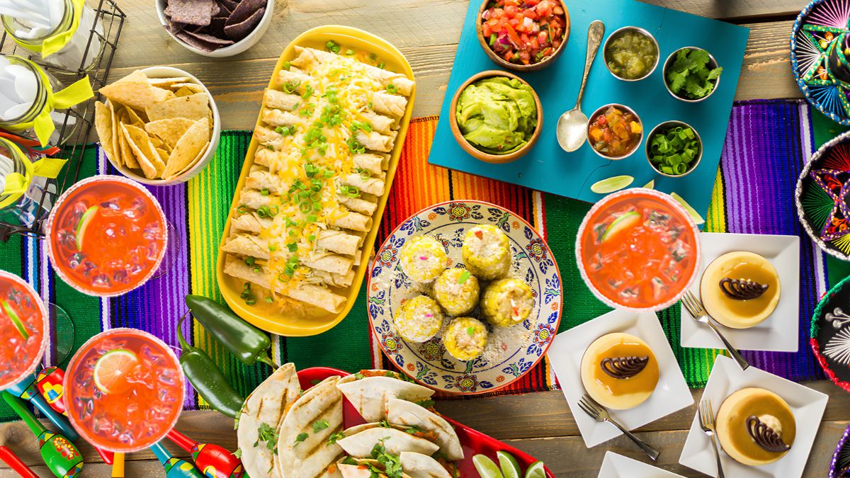 An overhead view of a colorful Mexican food spread featuring enchiladas, tacos, guacamole, salsa, flan, and drinks on a traditional striped table runner.