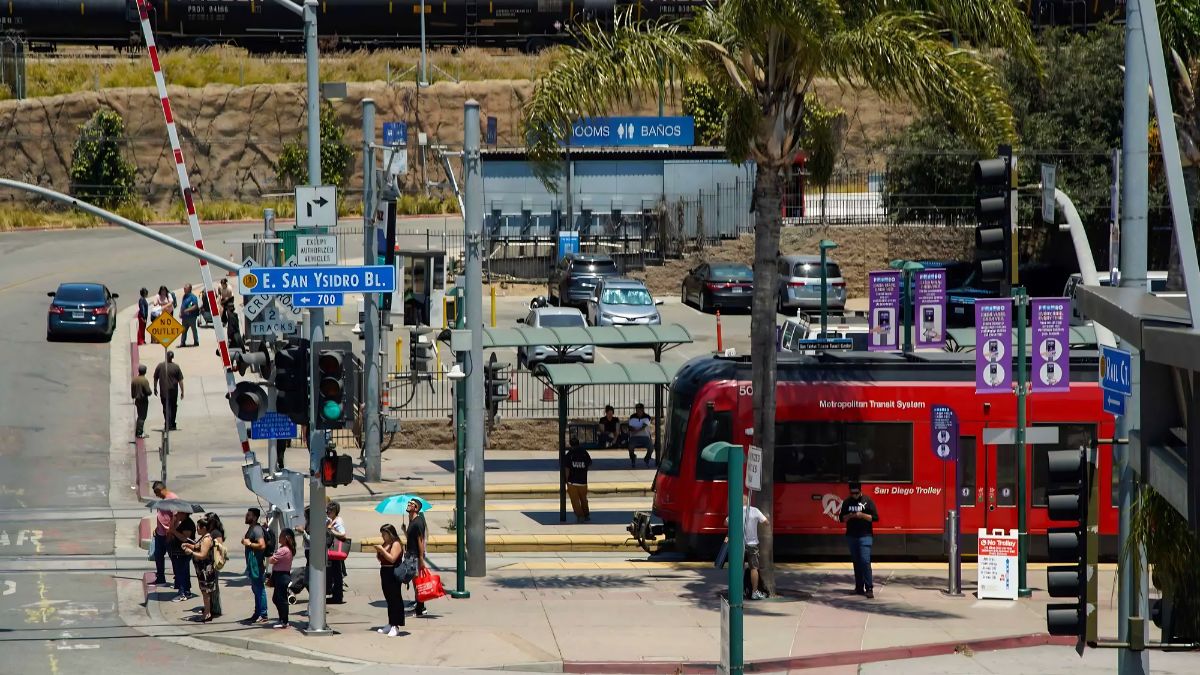 A San Ysidro transit station and street corner with a red trolley, pedestrians waiting, and street signs showing E. San Ysidro Blvd, illustrating multi-stop travel planning.