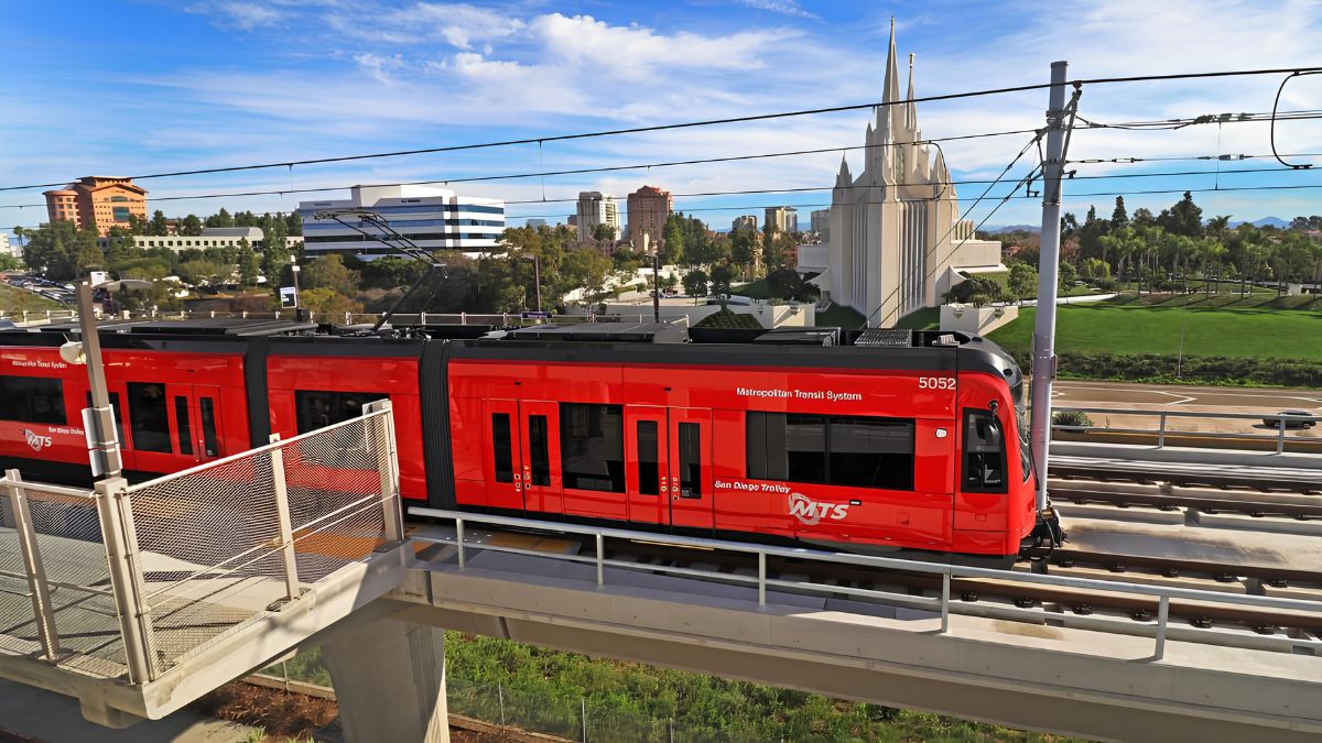 An elevated view of a red San Diego Trolley (MTS) train, number 5052, traveling on a concrete overpass. The train features the "Metropolitan Transit System" and "San Diego Trolley" logos. In the background, the prominent spires of a large white temple are visible, along with city buildings under a blue sky.