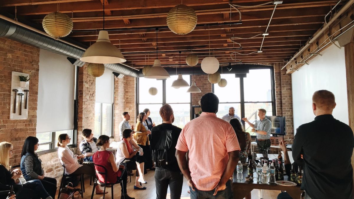 A group of people gathered inside a modern industrial venue with exposed brick walls, wooden beam ceilings, and hanging paper lanterns. Some attendees are seated on chairs, while others stand and listen to a speaker presenting near large windows.
