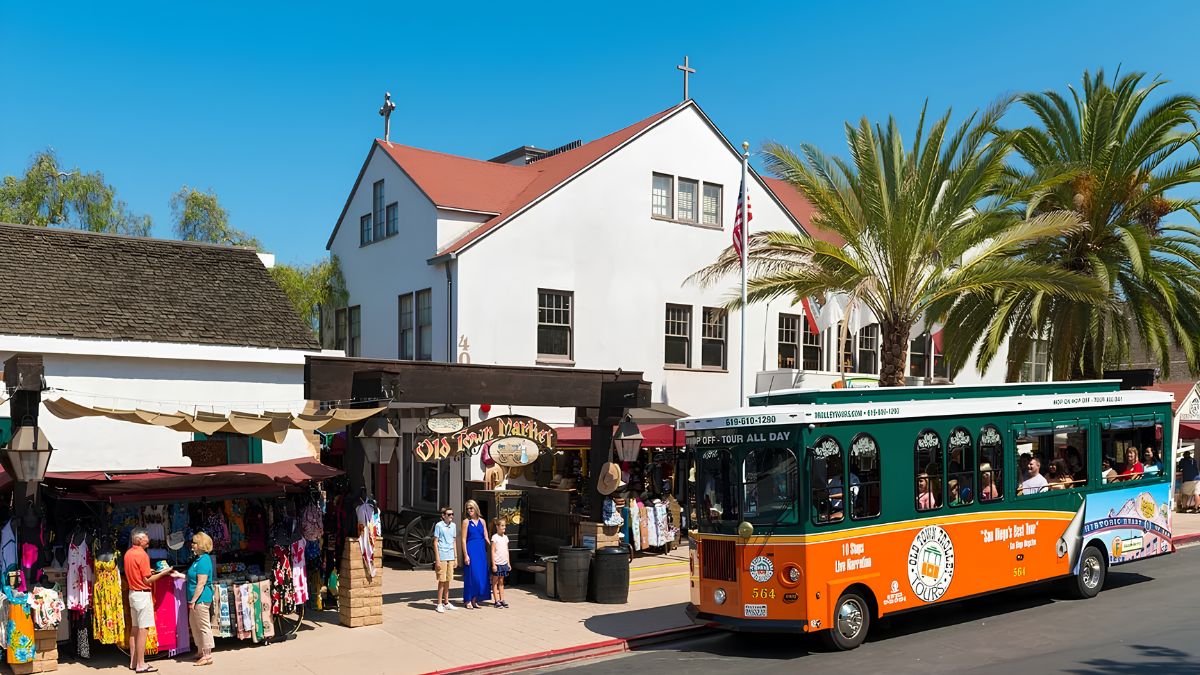 A green and orange sightseeing trolley driving past a historic building, market stalls, and a palm tree under a bright blue sky.