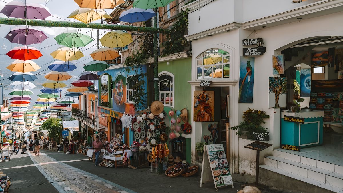 A vibrant sloping street lined with colorful shops, art galleries, and a canopy of hanging umbrellas overhead.