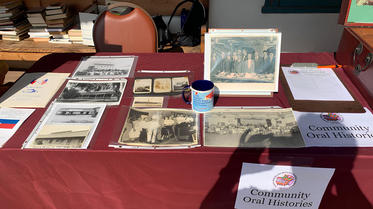 A table covered with a burgundy cloth displaying various black-and-white historic photographs, documents, a coffee mug, and a sign reading "Community Oral Histories".