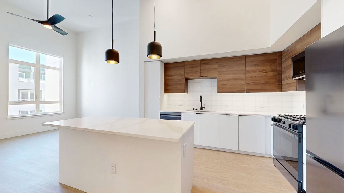 A modern, bright apartment kitchen with white countertops, wooden cabinets, and hanging pendant lights.
