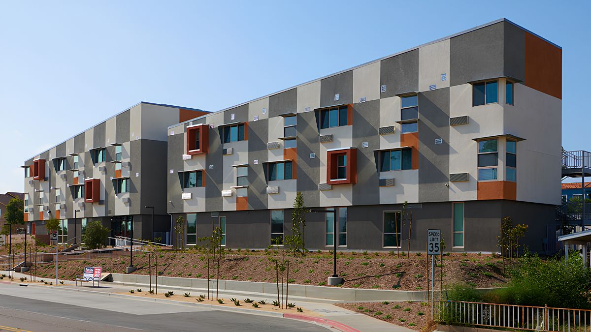 An exterior daytime photograph of a three-story modern apartment building in El Cajon, California. The facade has a distinctive patchwork pattern of grey, beige, and white stucco panels, accented by projecting, rust-colored window boxes. The building sits on a slope with new, drought-tolerant landscaping and terraced retaining walls. A sidewalk and street are in the foreground. A speed limit 35 sign is visible on the right.