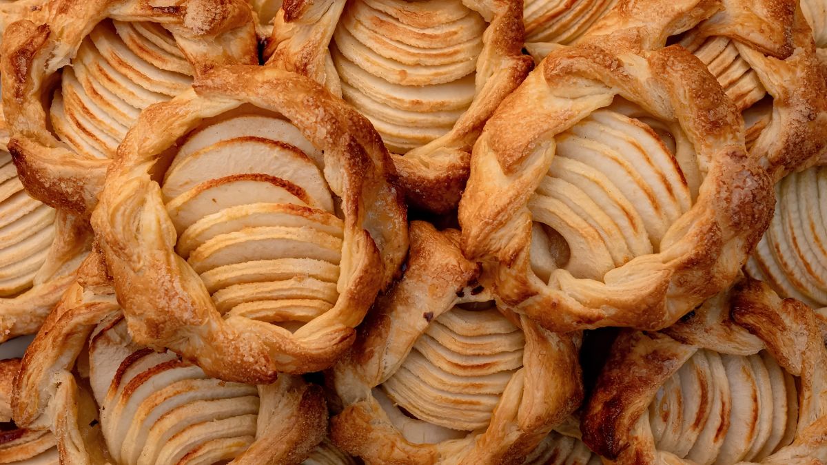 A close-up, overhead photograph of several freshly baked, golden-brown puff pastry apple tarts. Each tart features thinly sliced apples arranged in a fan or rosette pattern, dusted with sugar, and encased in a flaky, braided pastry crust. The tarts are arranged closely together, filling the frame.
