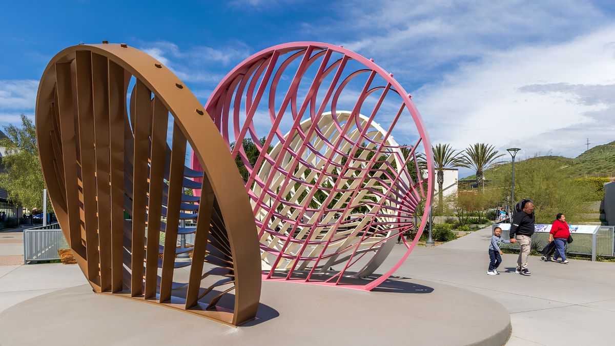 A large, modern outdoor sculpture in a sunlit plaza. It consists of two massive interlocking, curved metal structures. The left structure is solid, louvered, and bronze-colored. The right structure is a pink-colored lattice or mesh grid that forms a hollow sphere. On the right, a family (adult man, adult woman, and child) walks on a paved pathway near the artwork. Palm trees and a hillside are visible in the background under a blue sky with clouds.