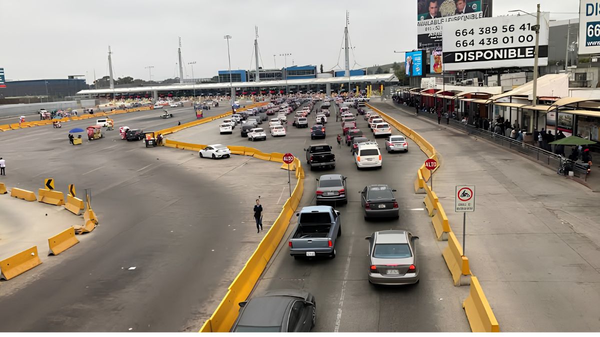 An elevated view of the busy San Ysidro Port of Entry vehicle crossing from Mexico to the US. Multiple lanes of cars and trucks are funneled between yellow concrete barriers towards the inspection facility in the background. A large billboard with Mexican phone numbers is on the right.