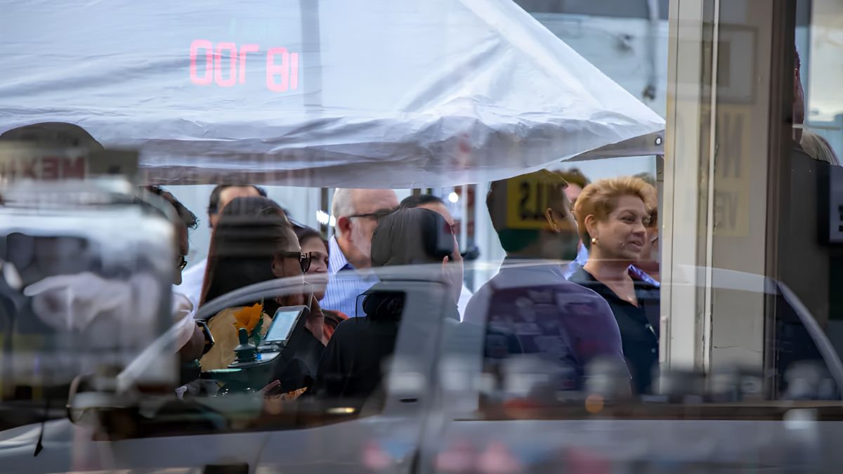 A candid outdoor shot looking through a street-side window at a small gathering of people, with a woman smiling in the foreground.