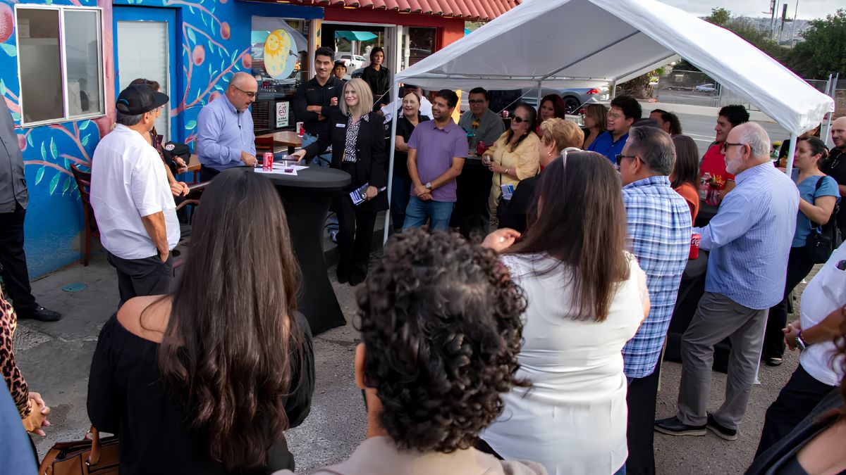 A crowd of people of varying ages and ethnicities gathered outdoors, facing a few speakers standing behind a small round table. The setting is an outdoor patio next to a building with a vibrant, colorful mural painted on its blue wall. A white pop-up canopy tent is set up to the side, and a street is visible in the background.