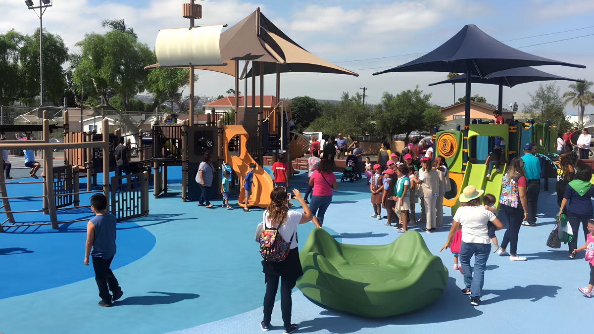 A wide, sunlit photograph capturing the grand opening of a vibrant community playground. Dozens of children and adults are enjoying the new, modern play structures with blue, green, and brown accents. A large group of young girls in matching uniforms and pink hats stand near a green, sculptural play piece. The ground is a blue soft-surfaced safety material. Palm trees and houses are visible in the background.