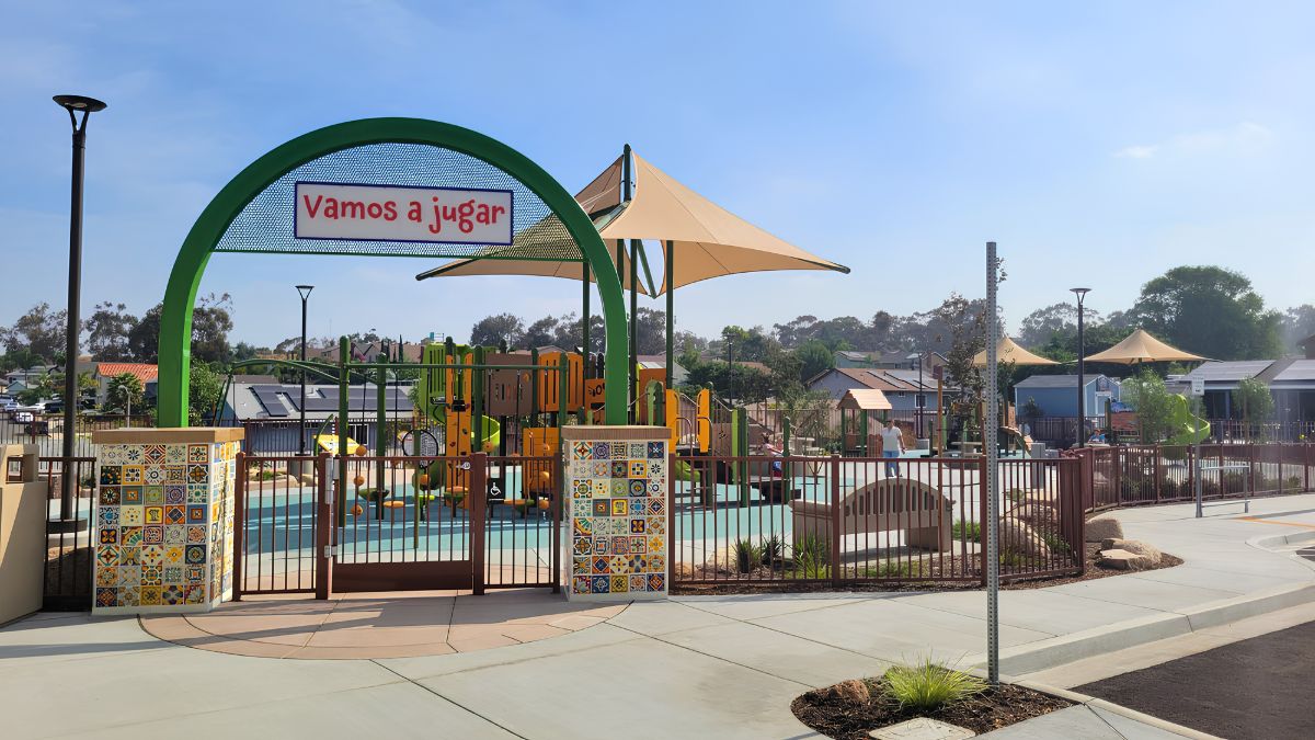 An eye-level shot of the entrance to a colorful community playground. A prominent green archway over a brown metal gate features a blue sign with the Spanish text "Vamos a Jugar." The gate is flanked by pillars decorated with intricate, colorful patterned tiles. Inside, modern yellow playground equipment is visible under a large tan shade sail, with housing and trees in the background under a clear sky.