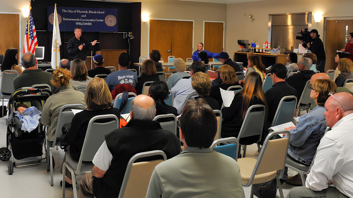 A medium-wide photograph taken from the back of a community meeting room. Dozens of attendees are seated in gray chairs, facing a speaker at a podium in the front. A banner behind the speaker reads "The City of Warwick, Rhode Island and Buttonwoods Community Center Welcomes." American and Rhode Island flags flank the banner. A baby stroller is visible in the left foreground, and a videographer is filming from the back right.