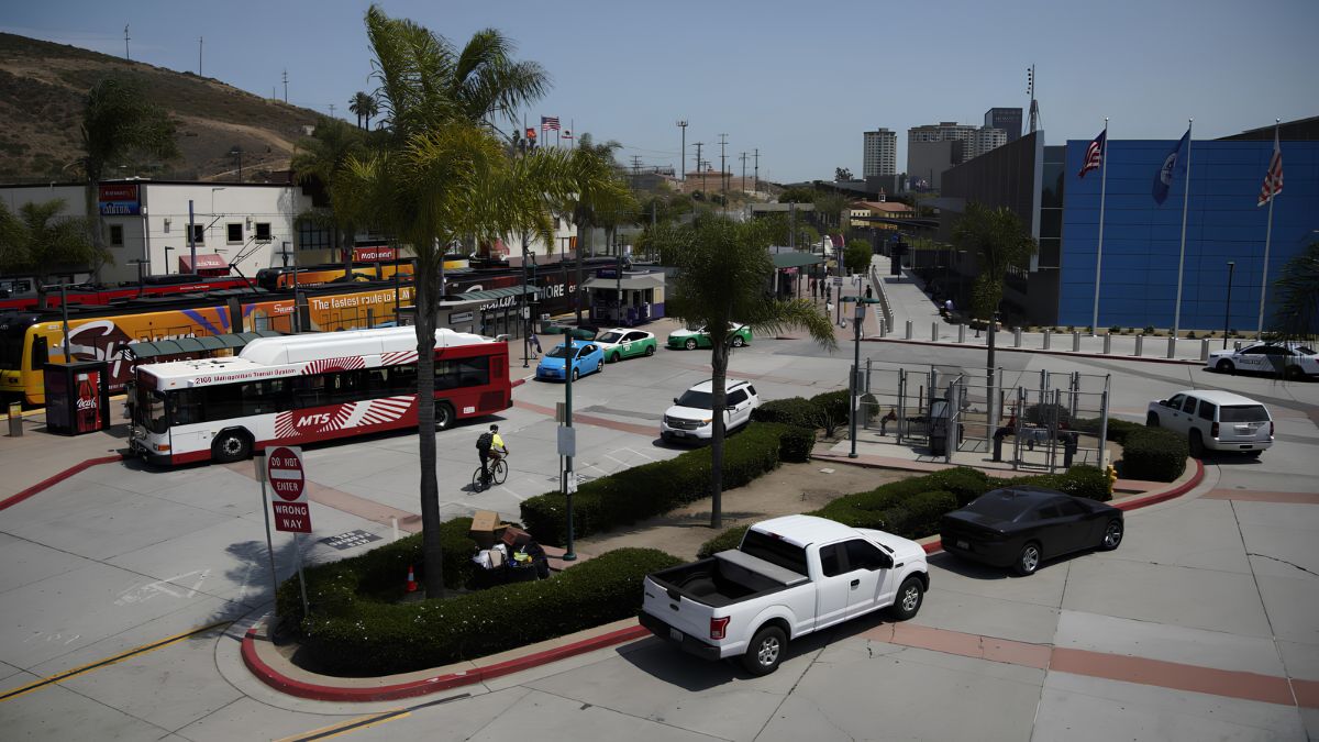 A high-angle, sunny photograph of the San Ysidro Transit Center, featuring a red Metropolitan Transit System (MTS) bus and yellow trolleys. In the foreground, a white Ford pickup truck is parked near green hedges. A pedestrian walkway leads to the U.S. port of entry buildings, which display American flags.