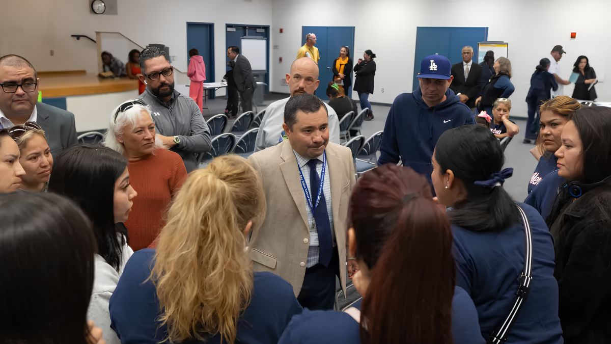 A man in a beige suit and blue tie stands at the center of a crowd of people, engaged in a serious discussion. The surrounding group includes women with blonde and dark hair, and men in varied attire, all focused on him.