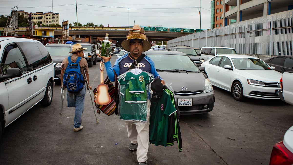 A street vendor in a sombrero walking between stopped cars, holding a stack of green Mexican soccer jerseys, a ukulele, and a bottle. Another person with a backpack walks ahead using crutches.