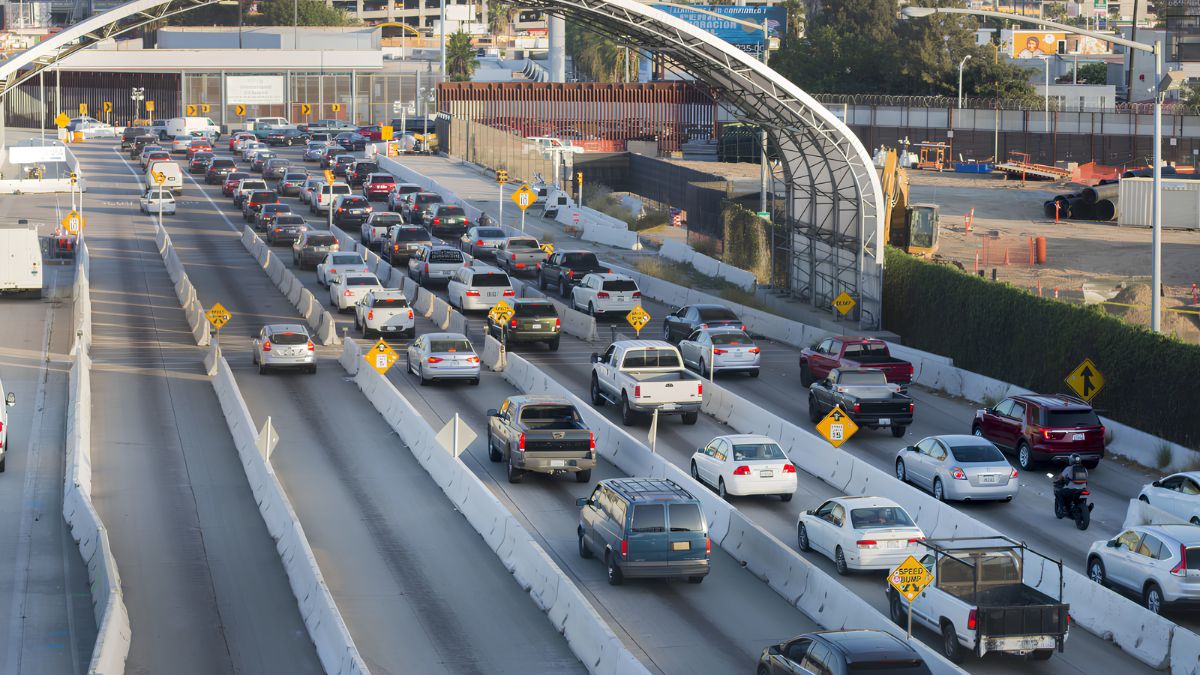 An elevated, high-angle view of heavy vehicular traffic waiting in multi-lane queues at a major border crossing, separated by concrete barriers, with infrastructure and signs visible in the background.