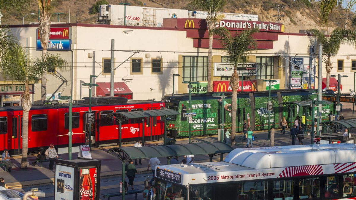 An elevated view of a busy outdoor transit plaza in San Ysidro. Prominent red and green San Diego Trolley trains are at the platform, with a large McDonald's sign on a building in the background. People are walking and waiting. Palm trees and commercial storefronts line the area under bright sunlight.