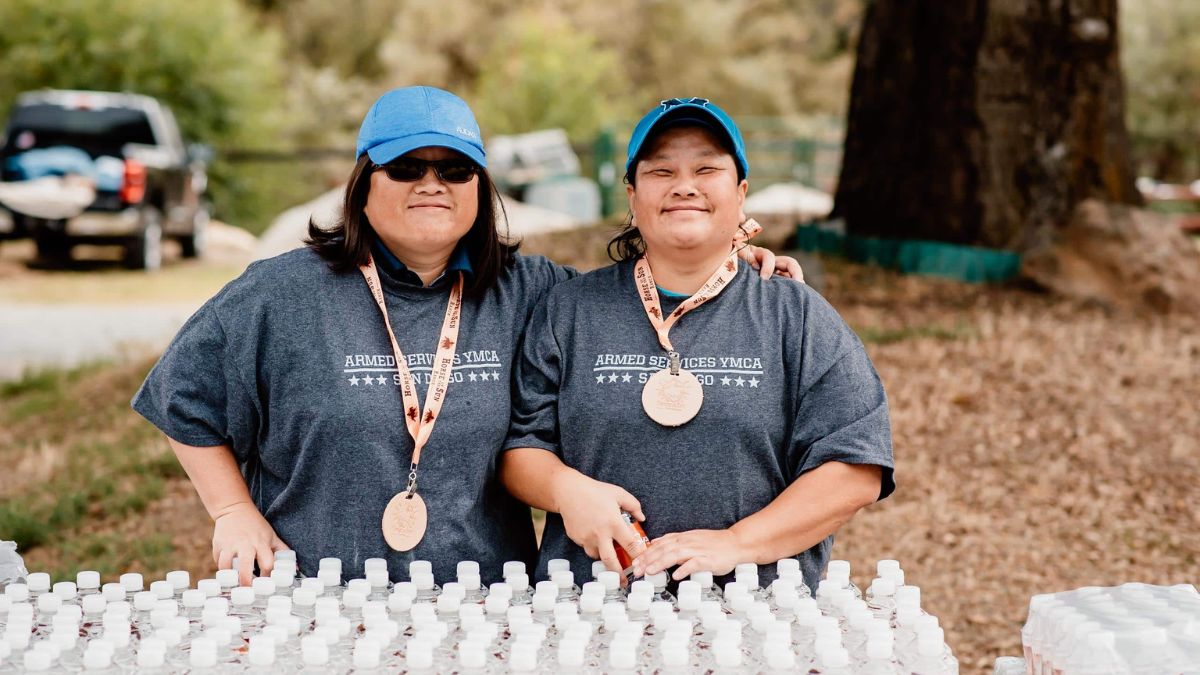 Two smiling volunteers wearing blue hats and matching grey "ARMED SERVICES YMCA" t-shirts stand behind a table lined with dozens of small bottled water containers during an outdoor community event.
