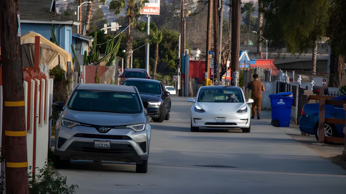 Cars driving and parked along a neighborhood street lined with houses and utility poles in San Ysidro
