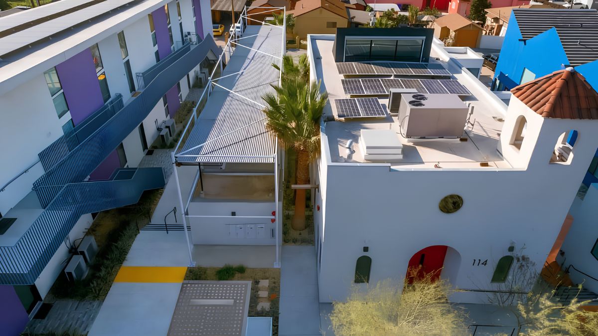 An aerial photograph of a modern apartment complex in Southern California, showing a central courtyard between a purple multi-story building and a white Spanish-style building with solar panels and a bell tower. The courtyard features a white metal trellis and a palm tree.