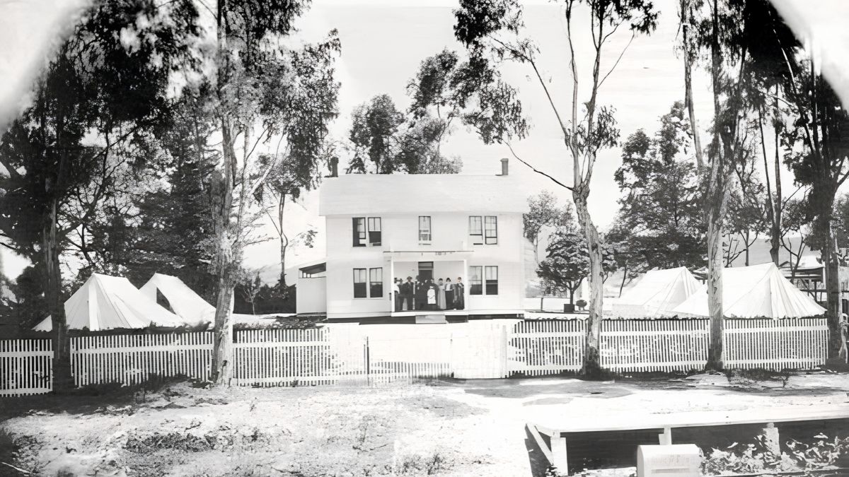 A vintage black-and-white archival photograph of a historic two-story house behind a white picket fence with tents and tall trees in San Ysidro.
