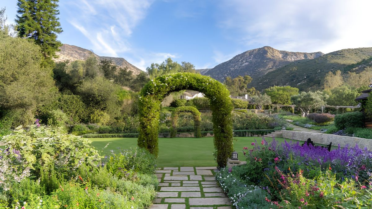 A lush green garden path leading under a trimmed topiary arch, surrounded by flowering plants, trees, and distant mountains in San Ysidro
