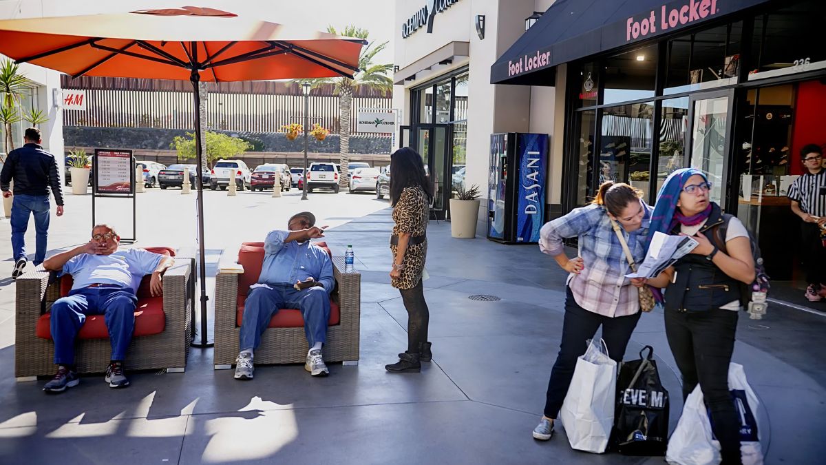 A daytime outdoor shopping plaza walkway. Two men relax in red armchairs under an umbrella on the left. In the foreground on the right, a woman in a hijab holds papers while another woman stands beside her. In the background are storefronts like Foot Locker and H&M, with cars parked further back.