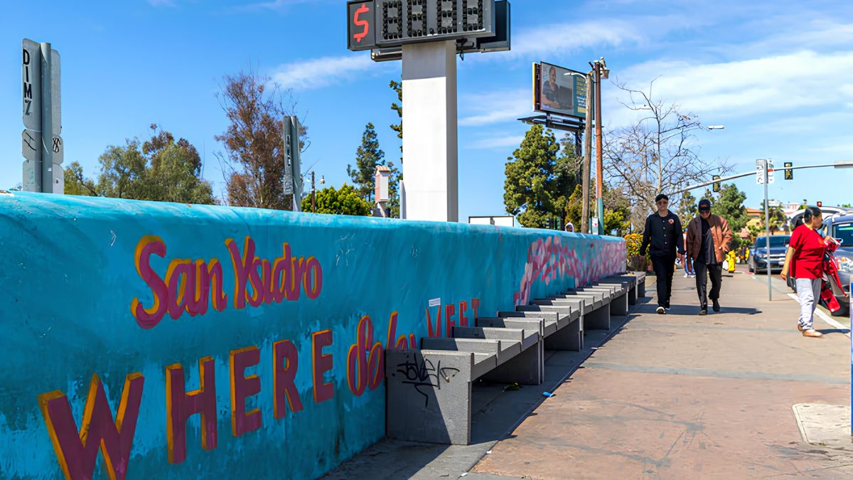 A colorful wall mural with text reading "San Ysidro" along a sidewalk with pedestrians walking by
