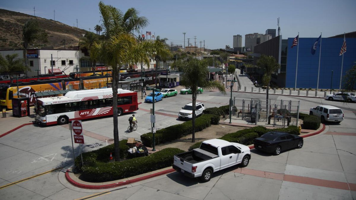 An elevated, sun-drenched photograph of a multi-modal transit hub. In the foreground, a red and white MTS bus is stopped, and a person on a bicycle rides nearby. Palm trees are dotted throughout the paved area. To the left, yellow and orange San Diego Trolley light rail vehicles are visible. Several cars and a white pickup truck are parked or driving through. In the background, a pedestrian bridge leads toward a large, modern blue building with flags, and beyond that, the urban landscape of a distant city rises against a hillside under a clear blue sky.
