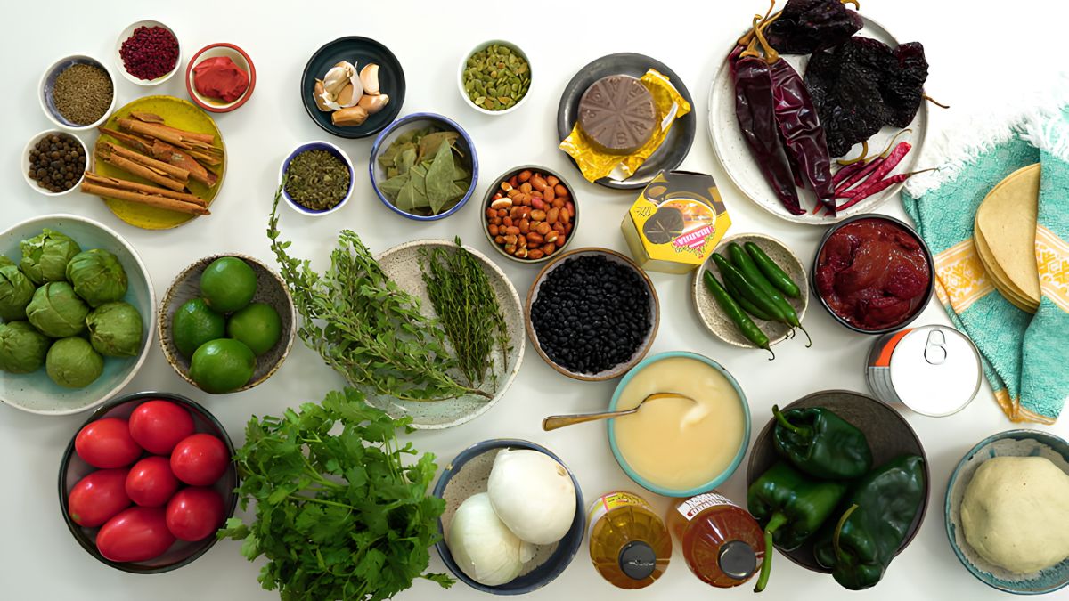 An overhead photograph displaying a wide array of raw Mexican ingredients for making mole Poblano arranged neatly on a white table. The ingredients include various dried and fresh chiles, spices, nuts, herbs, produce, and dough.