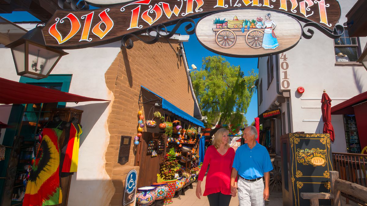 A sunny, wide-angle photograph of a senior couple walking hand-in-hand down a vibrant, cobblestone pathway in Old Town Market, San Diego. They are smiling and looking at each other. They are surrounded by colorful artisan shops, pottery, and traditional Mexican textiles. A large sign for "Old Town Market" is overhead.