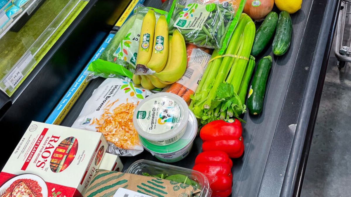 A grocery store checkout conveyor belt filled with fresh vegetables, bananas, and packaged food items.