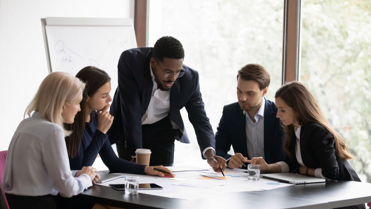 A diverse team of five business professionals in an office. An African American man in a suit leans over a table, pointing at documents, while his colleagues (one blonde woman, one brunette woman, two men in suits) sit and listen intently. A whiteboard is in the background.