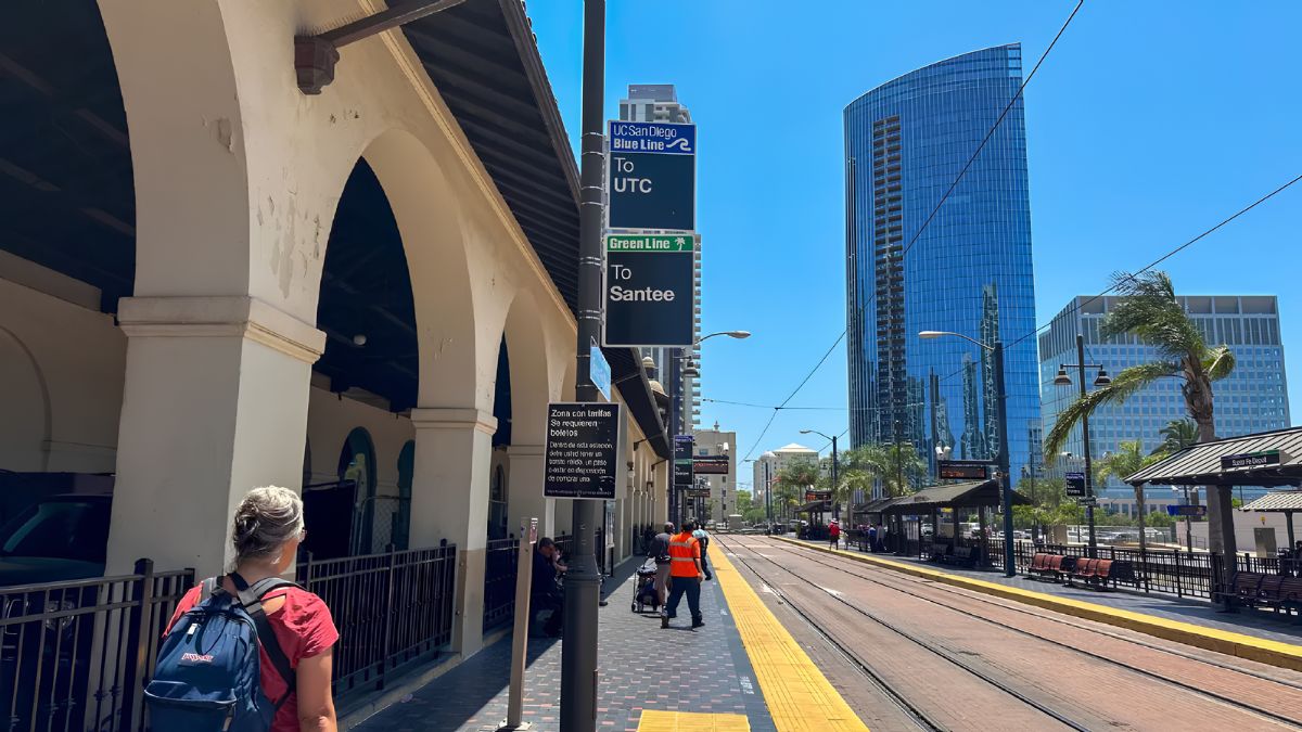Traveler standing on a San Diego Trolley station platform with transit line signage, railway tracks, and downtown skyscrapers in the background