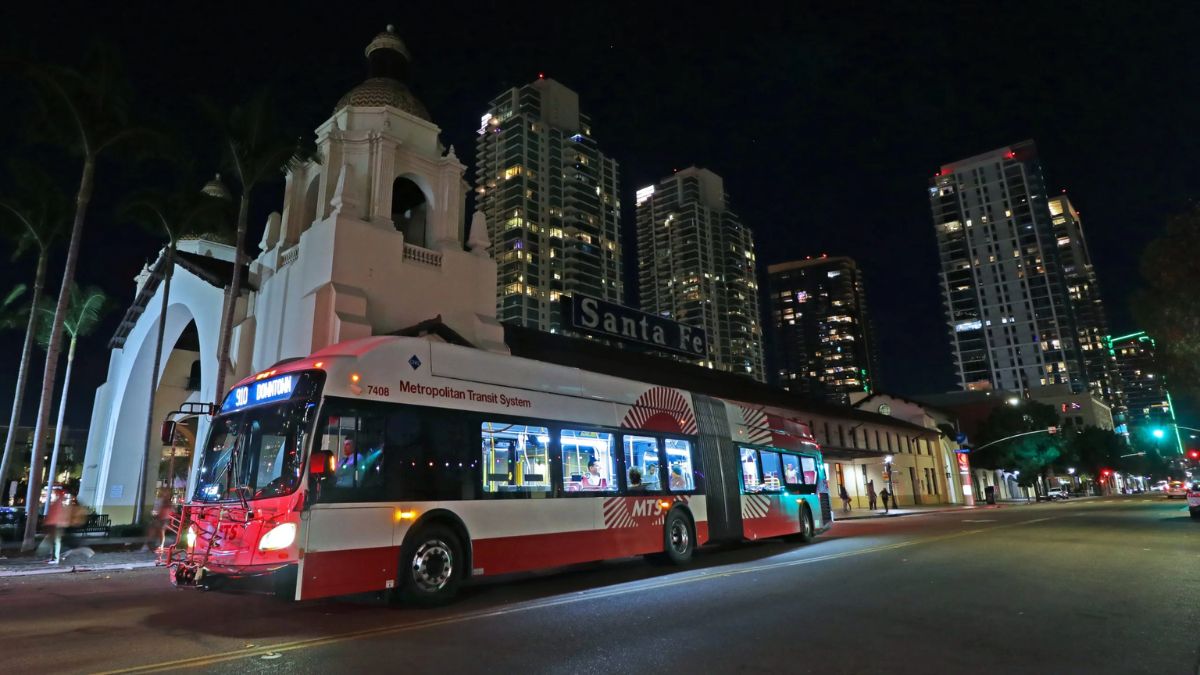 A San Diego Metropolitan Transit System (MTS) articulated bus operating at night near the Santa Fe station with downtown skyscrapers in the background.