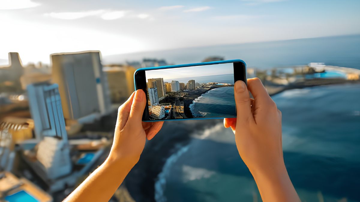 A traveler holding a smartphone to capture a coastal city view and ocean shoreline, contrasting with the theme of navigating a destination without digital devices.