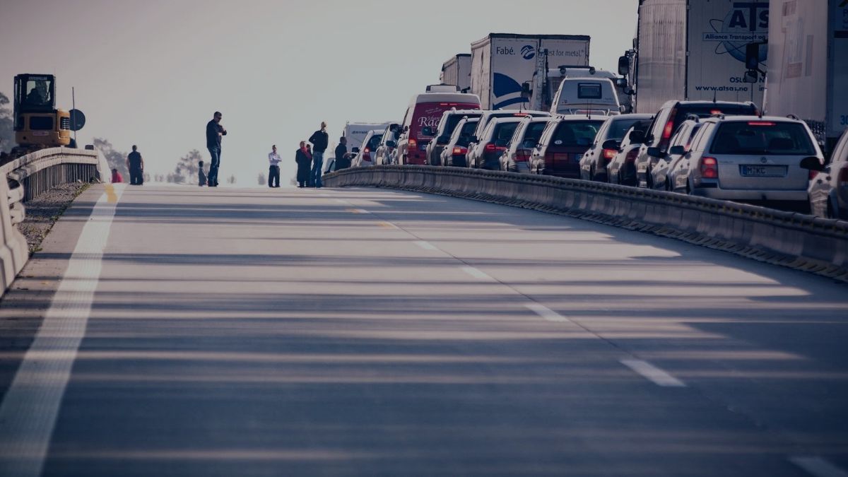 Heavy traffic congestion with cars and commercial trucks lined up on a highway, with pedestrians standing on the side.