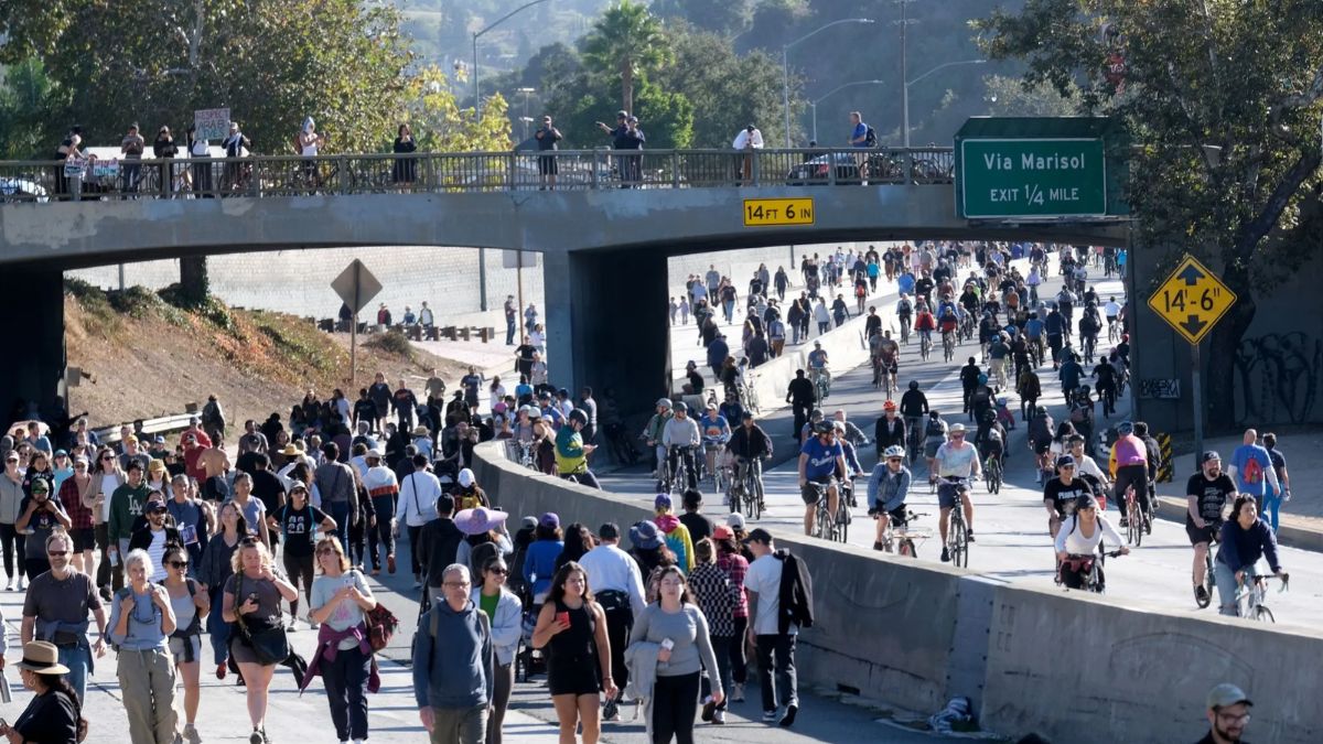 An elevated daytime photograph of a large crowd of people walking and cycling on a wide, open Los Angeles freeway, completely devoid of cars. A green highway sign reads "Via Marisol EXIT 1/4 MILE".