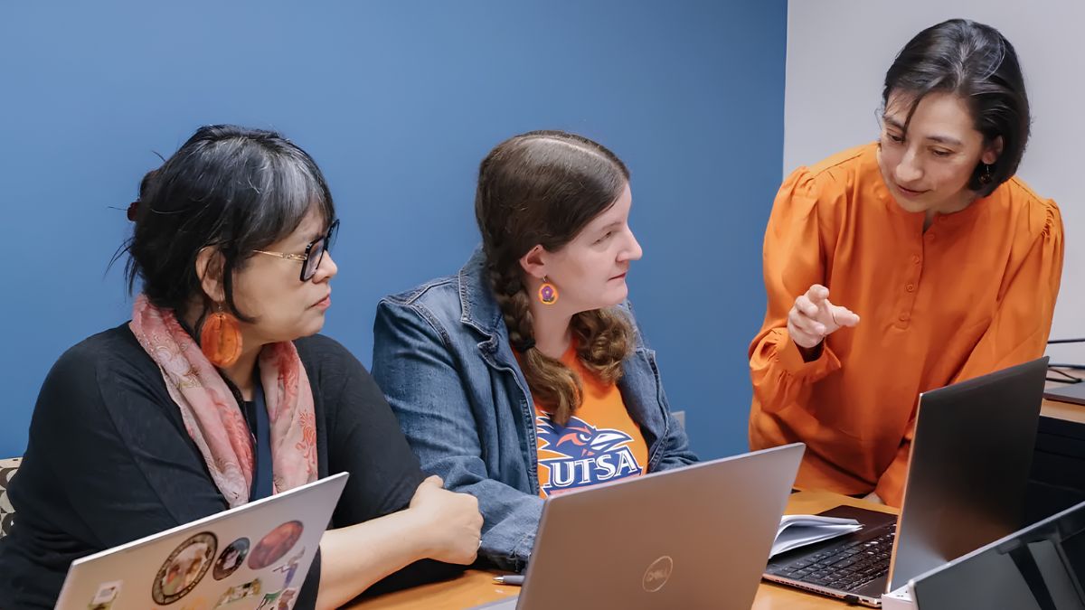 Three adults working together at a table with laptops while one instructor gestures and explains a digital task
